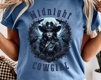 Può includere: T-shirt blu con una grafica di una cowgirl con un cappello, circondata da rose e corvi, con le parole "Midnight Cowgirl". Il design è in tonalità di nero e grigio.