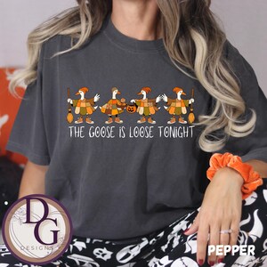 Könnte beinhalten: Dunkelgraues T-Shirt mit einer Reihe von Comic-Gänsen in Herbstkleidung, die Besen und einen Kürbis halten. Der Text "THE GOOSE IS LOOSE TONIGHT" ist darunter gedruckt. Das T-Shirt hat eine lockere Passform.
