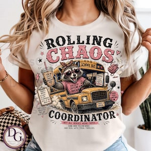May include: A cream-colored t-shirt featuring a graphic of a raccoon driving a yellow school bus. The text reads ROLLING CHAOS COORDINATOR, FUELED BY CAFFEINE & SARCASM, and includes details like a coffee cup, a clipboard, and school bus imagery.