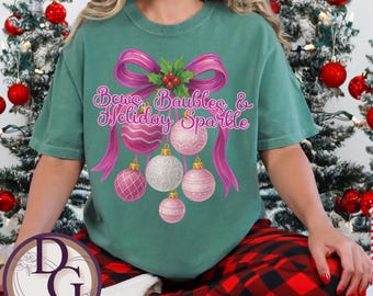 May include: Light green t-shirt with the text "Bows, Baubles & Holiday Sparkle" in a script font. The design features a pink bow, holly, and pink and white Christmas ornaments. The shirt is part of a holiday collection.