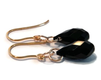 May include: A pair of drop earrings featuring faceted black teardrop-shaped beads suspended from gold-toned metal ear wires. Each earring consists of a single dark stone attached to a circular jump ring connected to a curved hook for the ear.