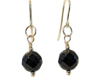 May include: A pair of gold-colored dangle earrings. Each earring features a faceted, black, round bead suspended from a gold-colored hook. The beads have a shiny, polished appearance, reflecting light.