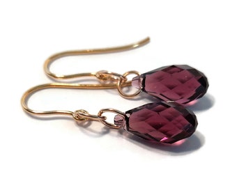 May include: A pair of drop earrings featuring faceted, teardrop-shaped purple crystals suspended from gold-toned metal ear wires. Each crystal is attached to the hook by a small circular jump ring, creating a simple and elegant dangling design.