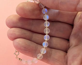 May include: A white iridescent beaded bracelet with a gold clasp. The bracelet is made of round beads and is strung on a gold chain.