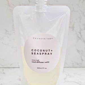 May include: Transparent plastic pouch with a white cap, filled with a pale yellow liquid. The pouch features a white label with the text "COCONUT + SEASPRAY" and "scented reed diffuser refill". The pouch contains 200ml of liquid.