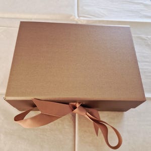 May include: A bronze-coloured gift box with a matching ribbon tied in a bow. The box has a textured surface and a simple, elegant design. The ribbon adds a decorative touch.