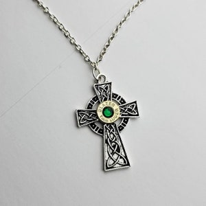 May include: A silver chain necklace with a Celtic cross pendant. The cross is decorated with a silver bullet casing with a green gem in the center. The bullet casing is inscribed with "Blazer 9mm Luger".