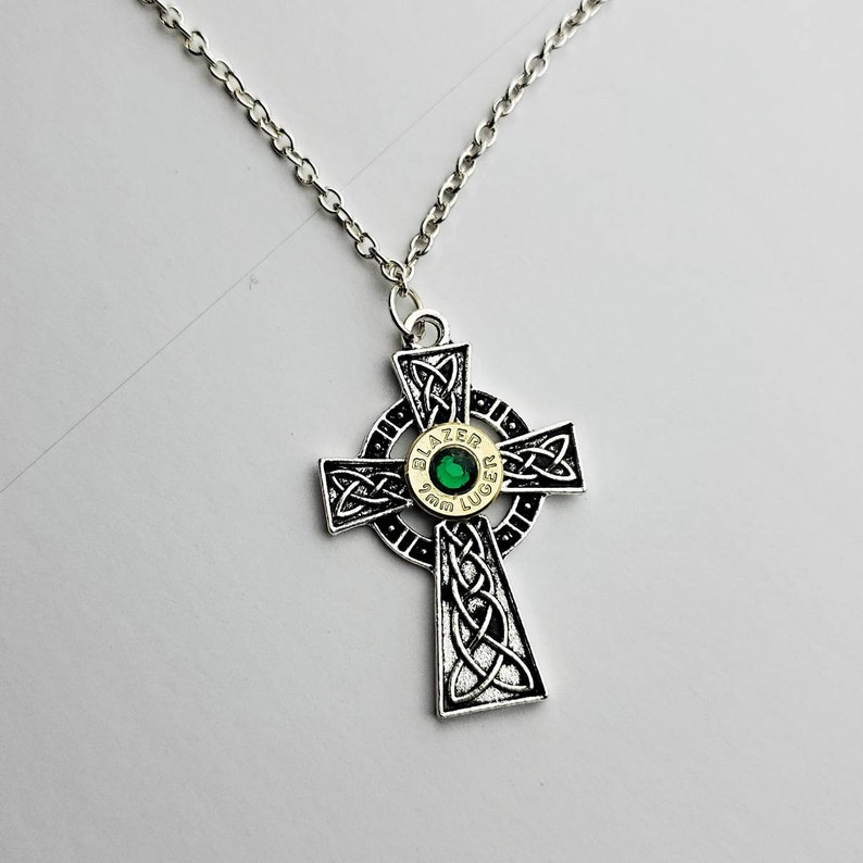 May include: A silver chain necklace with a Celtic cross pendant. The cross is decorated with a silver bullet casing with a green gem in the center. The bullet casing is inscribed with "Blazer 9mm Luger".