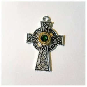 May include: Silver Celtic cross pendant with a gold-colored bullet casing in the center. The bullet casing has a green gemstone in the center and the words "9mm Luger" inscribed around the rim.