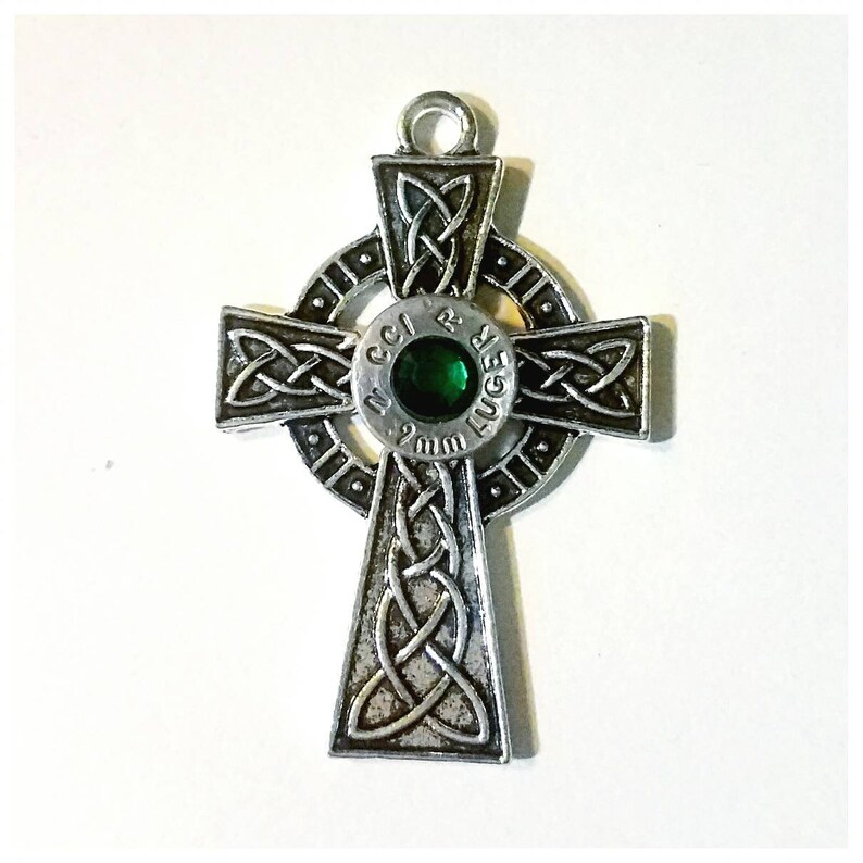 May include: A silver Celtic cross pendant with a 9mm Luger bullet casing in the center. The bullet casing has a green gemstone set in it.