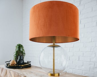 Op de afbeelding: Een gouden tafellamp met een heldere glazen bol als basis en een oranje fluwelen lampenkap. De lamp staat op een houten tafel met een chevron-patroon.