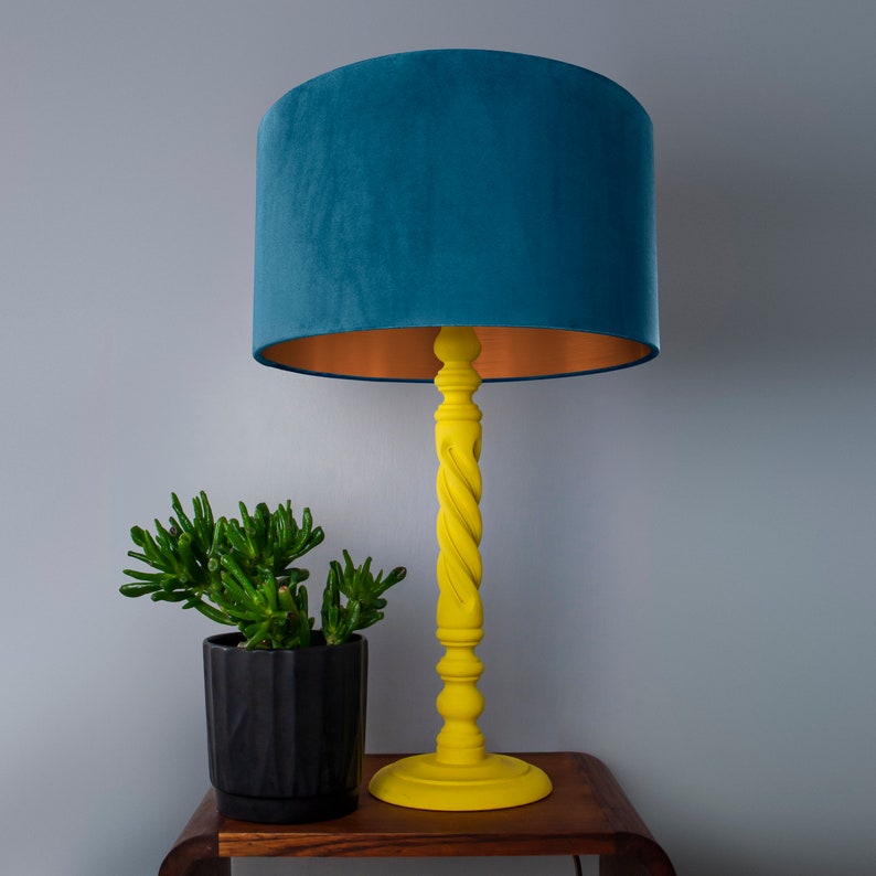 Puede incluir: Una l&aacute;mpara de mesa amarilla con una pantalla de terciopelo azul verdoso y un ribete de cobre. La l&aacute;mpara tiene una base de madera torcida y est&aacute; sobre una mesa de madera.