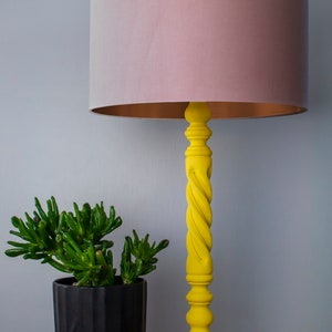 May include: A yellow table lamp with a twisted column and a round base. The lampshade is a soft pink color with a gold interior. A small green plant in a black pot sits next to the lamp. The lamp is on a wooden table.