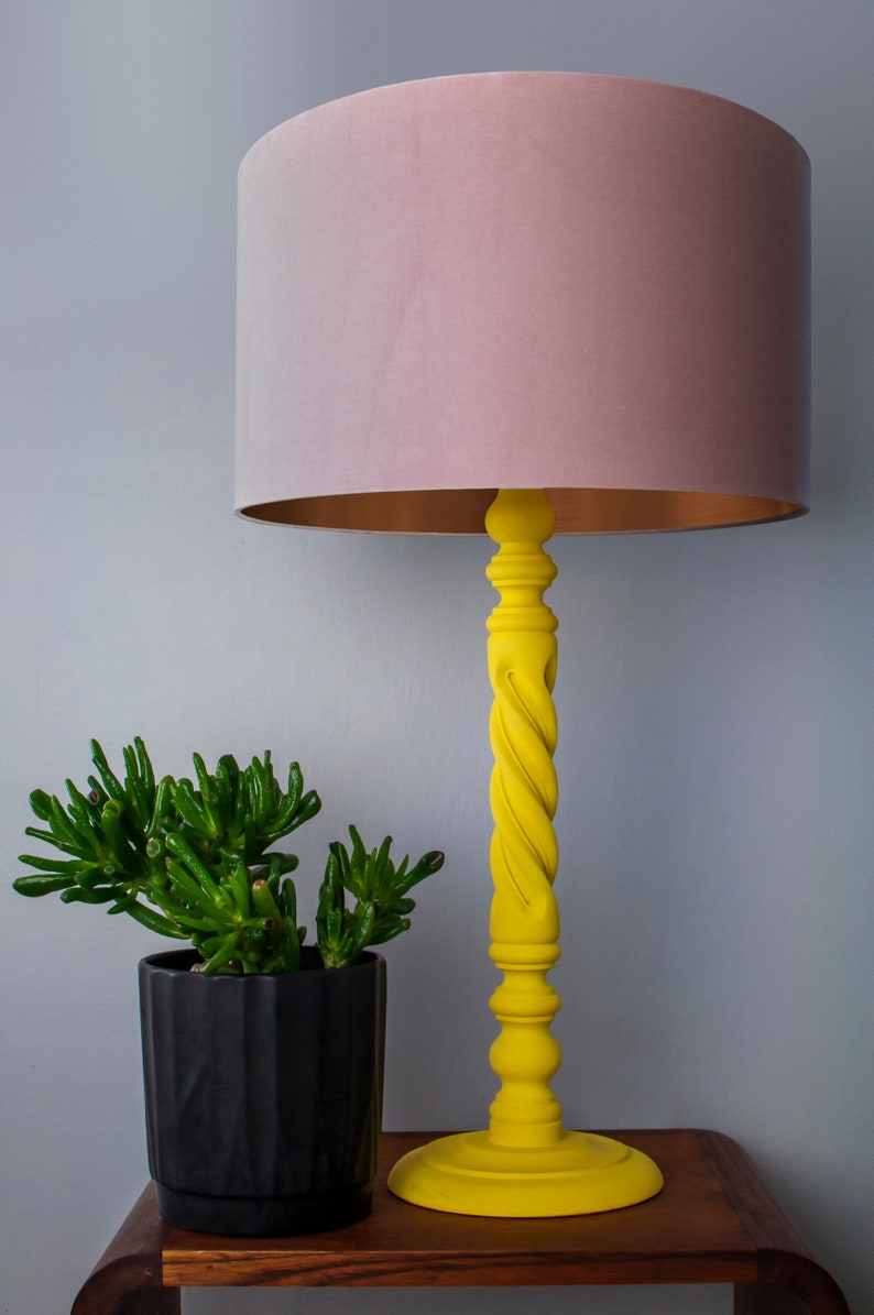 May include: A yellow table lamp with a twisted column and a round base. The lampshade is a soft pink color with a gold interior. A small green plant in a black pot sits next to the lamp. The lamp is on a wooden table.