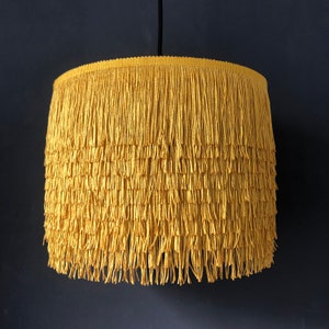 May include: A yellow fringe lampshade with a simple design. The lampshade is made of a soft, textured fabric and has a delicate fringe around the bottom.