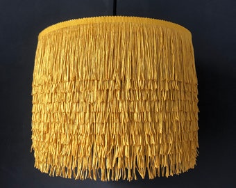 Op de afbeelding: Een gele lampenkap met franjes en een eenvoudig ontwerp. De lampenkap is gemaakt van een zachte, gestructureerde stof en heeft een delicate franje aan de onderkant.