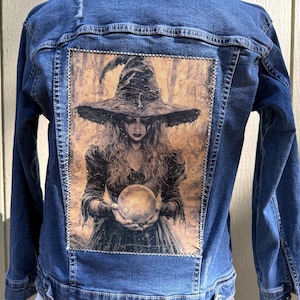 May include: Blue denim jacket with a large, rectangular patch on the back featuring a detailed illustration of a witch holding a crystal ball. The witch wears a large hat and dark clothing. The jacket has a classic design with button closures and pockets.