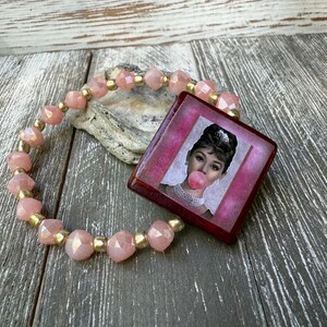 Könnte beinhalten: Ein rosa Perlenarmband mit goldenen Akzenten und einem quadratischen Anhänger mit einem Bild von Audrey Hepburn, die eine Kaugummiblase bläst. Der Anhänger hat einen rosa Rand und einen dunkelroten Rahmen.