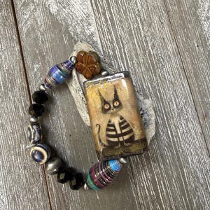 May include: A stretch bracelet featuring a rectangular focal piece with a graphic illustration of a cat with large eyes and striped fur. The bracelet includes assorted beads in black, blue, and brown, including cylindrical paper beads and a flower-shaped bead.