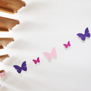 Può includere: Una ghirlanda di farfalle di carta in tonalità rosa e viola, appesa a un muro bianco.