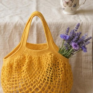 Puede incluir: Un bolso tote tejido a ganchillo de color amarillo mostaza con dos asas. El bolso tiene un diseño de red y está lleno de un objeto invisible. Un ramo de flores moradas y una taza con temática de mariposas están cerca.