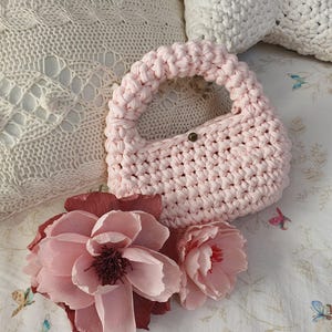 以下が含まれることがあります： ボタン留めのピンクのクロシェバッグ。バッグは白とピンクの模様の布の上に置かれ、ピンクの紙の花が2つあります。