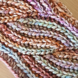 Peut inclure: Gros plan sur une écharpe en crochet multicolore avec un motif texturé. L'écharpe est réalisée avec des fils de différentes nuances de rose, orange, bleu et blanc.