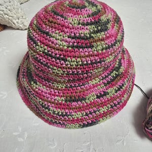 Puede incluir: Un sombrero de cubo de ganchillo en rosa, verde, marrón y gris. El sombrero está hecho con una variedad de hilos de diferentes colores.