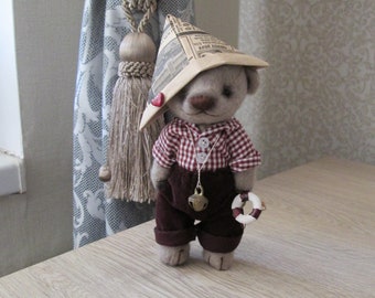 Puede incluir: Un oso de peluche marrón que lleva una camisa a cuadros rojos y blancos, pantalones marrones y un sombrero de papel. El oso sostiene un salvavidas blanco y una campana dorada.