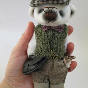 May include: A white teddy bear wearing a green sweater vest, brown bow tie, and a plaid newsboy cap. The bear is holding a brown leather briefcase and has a gold dollar sign charm attached to its vest.