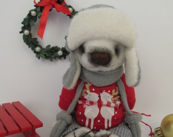 May include: A gray plush toy wearing a white fur hat, red sweater with reindeer design, and gray pants. The toy is sitting on a white surface with a red sled and a gold ornament in the background.