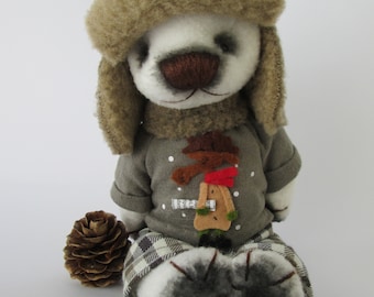 Puede incluir: Un oso de peluche blanco con un gorro de piel marrón con una estrella roja, un suéter gris con un diseño de muñeco de nieve y pantalones a cuadros. El oso está sentado sobre una superficie blanca.