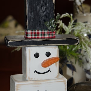 Puede incluir: Una decoraci&oacute;n de mu&ntilde;eco de nieve de madera con un sombrero de copa negro, una cinta de cuadros rojos y verdes y una cara blanca con ojos negros, una nariz naranja y una sonrisa negra. El mu&ntilde;eco de nieve est&aacute; hecho de tres bloques de madera apilados.