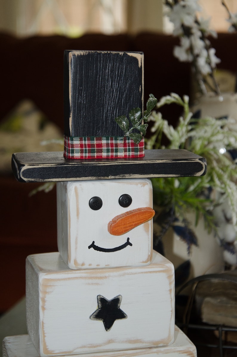 Puede incluir: Una decoraci&oacute;n de mu&ntilde;eco de nieve de madera con un sombrero de copa negro, una cinta de cuadros rojos y verdes y una cara blanca con ojos negros, una nariz naranja y una sonrisa negra. El mu&ntilde;eco de nieve est&aacute; hecho de tres bloques de madera apilados.