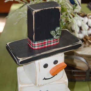 Puede incluir: Una decoraci&oacute;n de mu&ntilde;eco de nieve de madera con un sombrero de copa negro, un cuerpo blanco y una nariz naranja. El mu&ntilde;eco de nieve tiene ojos negros y una boca negra.