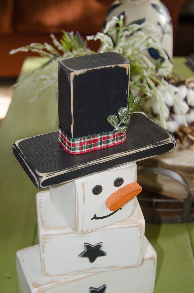 Puede incluir: Una decoraci&oacute;n de mu&ntilde;eco de nieve de madera con un sombrero de copa negro, un cuerpo blanco y una nariz naranja. El mu&ntilde;eco de nieve tiene ojos negros y una boca negra.