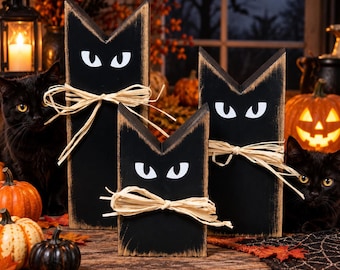 May include: Three black wooden cat-shaped decorations with pointed ears, white painted eyes, and natural raffia bows tied around the center. The wood edges feature a distressed, worn finish, and the figures stand in varying heights.