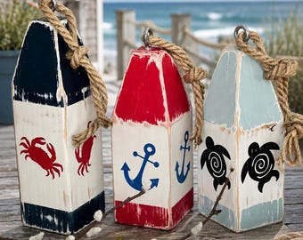 Peut inclure: Trois bouées de homard décoratives en bois avec des finitions de peinture usée. La bouée de gauche est bleu marine et blanche avec un crabe rouge, celle du centre est rouge et blanche avec des ancres bleues, et celle de droite est bleu clair.