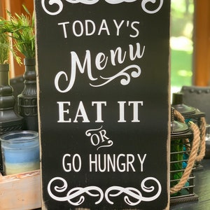 May include: Black wooden sign with white text that reads "Today's Menu Eat It Or Go Hungry". The sign has decorative flourishes at the top and bottom.