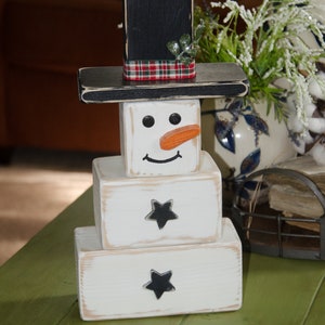 Puede incluir: Una decoraci&oacute;n de mu&ntilde;eco de nieve de madera con un sombrero negro, un cuerpo blanco y detalles de estrellas negras. El mu&ntilde;eco de nieve tiene una cinta de cuadros rojos y negros alrededor del sombrero y una nariz de zanahoria.