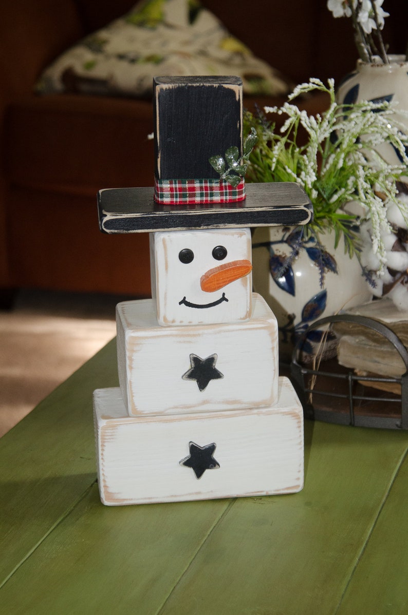 Puede incluir: Una decoraci&oacute;n de mu&ntilde;eco de nieve de madera con un sombrero negro, un cuerpo blanco y detalles de estrellas negras. El mu&ntilde;eco de nieve tiene una cinta de cuadros rojos y negros alrededor del sombrero y una nariz de zanahoria.