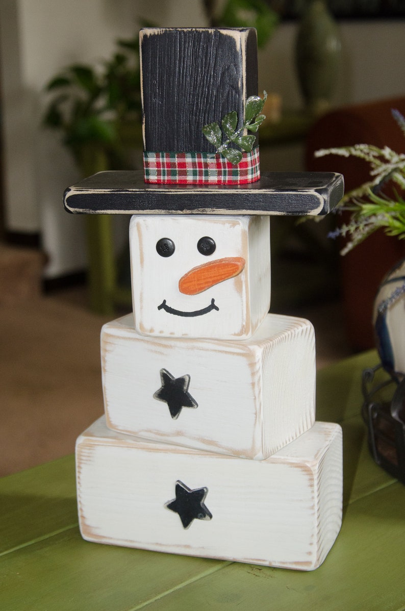 Puede incluir: Una decoraci&oacute;n de mu&ntilde;eco de nieve de madera con un sombrero de copa negro, una cinta de cuadros rojos y blancos y hojas verdes. El mu&ntilde;eco de nieve tiene una cara pintada de blanco con ojos negros, una sonrisa negra y una nariz de zanahoria naranja. El mu&ntilde;eco de nieve est&aacute; hecho de bloques de madera apilados con estrellas negras en los dos bloques inferiores.