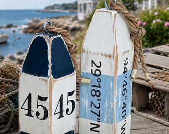 May include: Two wooden lobster buoys with distressed paint finishes. The smaller buoy is navy blue and white with the number 45 printed on it. The larger buoy is white with a light blue stripe and the coordinates 29°18'27" N 94°46'40" W.