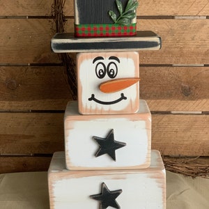 Puede incluir: Una decoraci&oacute;n de mu&ntilde;eco de nieve de madera con un sombrero de copa negro, una ramita verde y una cinta de cuadros rojos y verdes. El mu&ntilde;eco de nieve tiene una cara blanca con ojos negros, una nariz de zanahoria y una sonrisa negra. El mu&ntilde;eco de nieve est&aacute; hecho de tres bloques apilados, con estrellas negras en los dos bloques inferiores.