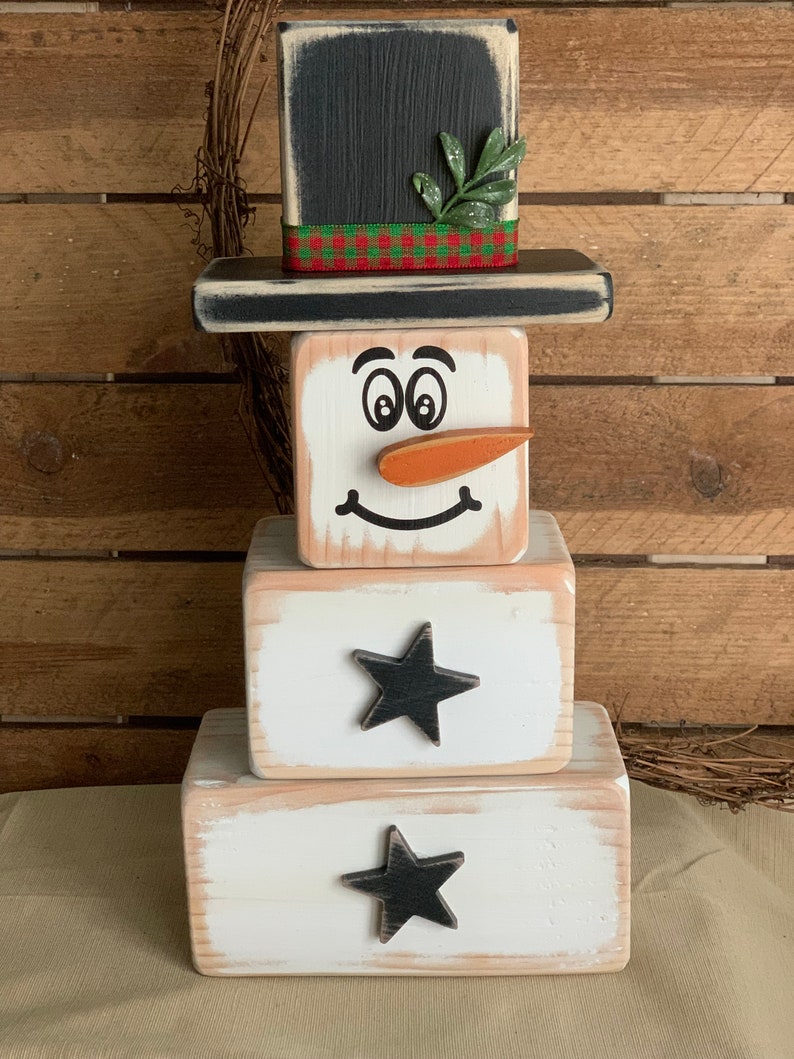 Puede incluir: Una decoraci&oacute;n de mu&ntilde;eco de nieve de madera con un sombrero de copa negro, una ramita verde y una cinta de cuadros rojos y verdes. El mu&ntilde;eco de nieve tiene una cara blanca con ojos negros, una nariz de zanahoria y una sonrisa negra. El mu&ntilde;eco de nieve est&aacute; hecho de tres bloques apilados, con estrellas negras en los dos bloques inferiores.