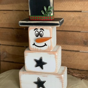 Puede incluir: Una figura de mu&ntilde;eco de nieve de madera con un sombrero de copa negro, una ramita verde y una cinta de cuadros rojos y verdes. El mu&ntilde;eco de nieve tiene una cara blanca con ojos negros, una nariz de zanahoria y una boca sonriente. El mu&ntilde;eco de nieve est&aacute; hecho de tres bloques apilados, con los dos bloques inferiores decorados con estrellas negras.
