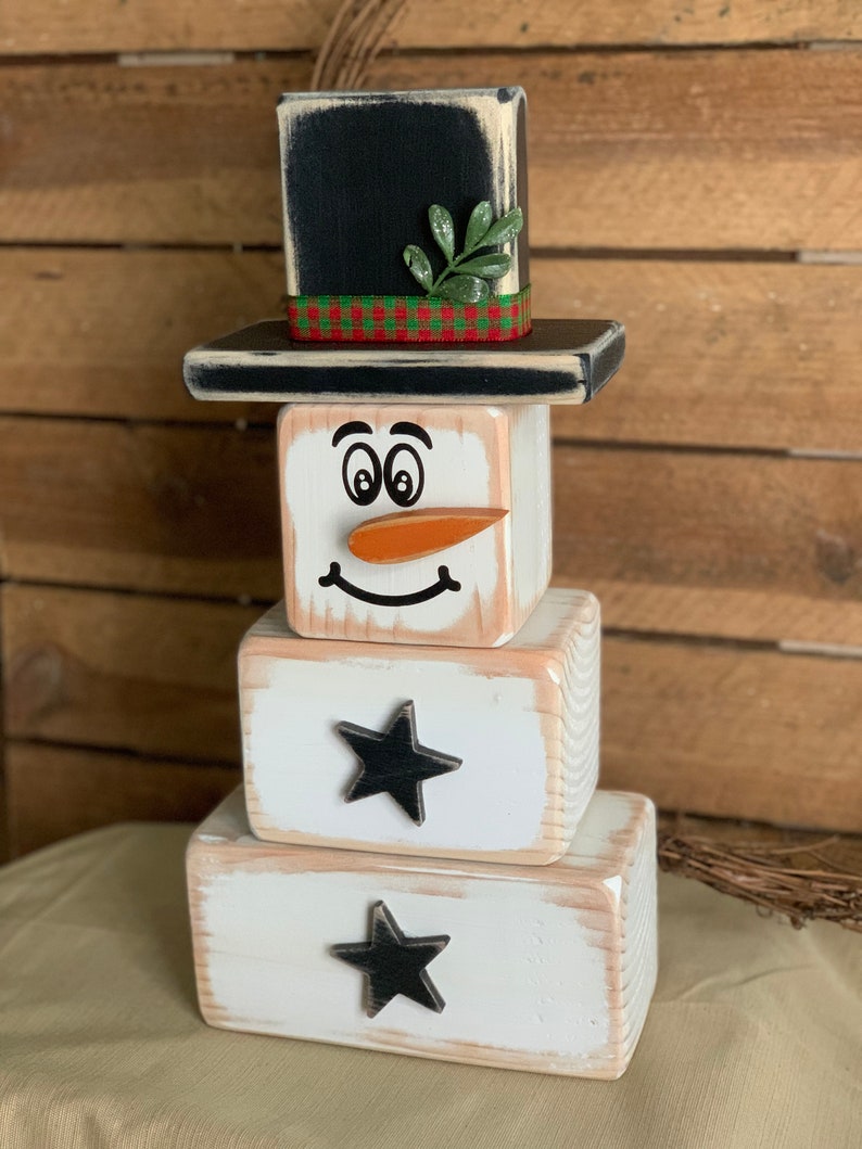 Puede incluir: Una figura de mu&ntilde;eco de nieve de madera con un sombrero de copa negro, una ramita verde y una cinta de cuadros rojos y verdes. El mu&ntilde;eco de nieve tiene una cara blanca con ojos negros, una nariz de zanahoria y una boca sonriente. El mu&ntilde;eco de nieve est&aacute; hecho de tres bloques apilados, con los dos bloques inferiores decorados con estrellas negras.