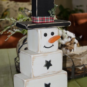 Puede incluir: Una decoraci&oacute;n de mu&ntilde;eco de nieve de madera con un sombrero de copa negro, una cinta a cuadros y una ramita verde. El mu&ntilde;eco de nieve est&aacute; hecho de tres bloques de madera blancos con ojos pintados de negro, una sonrisa pintada de negro y una nariz pintada de naranja. Los dos bloques inferiores tienen estrellas pintadas de negro.
