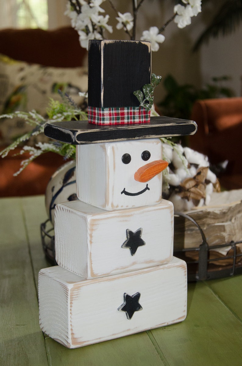 Puede incluir: Una decoraci&oacute;n de mu&ntilde;eco de nieve de madera con un sombrero de copa negro, una cinta a cuadros y una ramita verde. El mu&ntilde;eco de nieve est&aacute; hecho de tres bloques de madera blancos con ojos pintados de negro, una sonrisa pintada de negro y una nariz pintada de naranja. Los dos bloques inferiores tienen estrellas pintadas de negro.