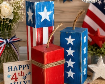 May include: Three rectangular wooden blocks painted in red, white, and blue with star motifs and jute rope accents. A small rectangular sign reads Happy 4th of July with blue text and a red number four. The blocks feature a distressed finish.